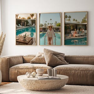 May include: Three framed art prints depicting scenes by a swimming pool. One shows a table with a champagne bucket, another a person in a white outfit, and the third a group of people. The prints are above a brown sofa and a stone coffee table.