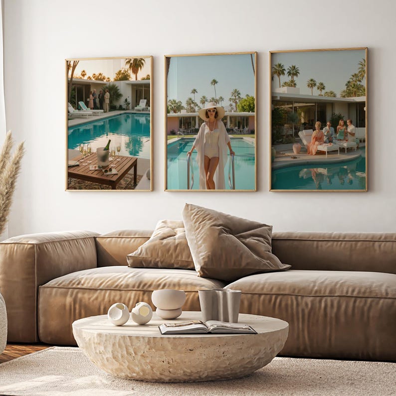 May include: Three framed art prints depicting scenes by a swimming pool. One shows a table with a champagne bucket, another a person in a white outfit, and the third a group of people. The prints are above a brown sofa and a stone coffee table.