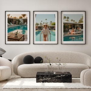 May include: Three framed art prints depicting poolside scenes. One shows a table with champagne, another a person in a white outfit by a pool, and the third, people lounging by a pool. The prints are displayed above a cream-colored sofa.