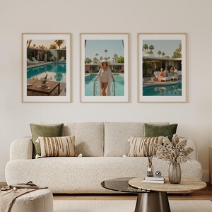 May include: Three framed art prints depicting a retro poolside scene with a turquoise pool, palm trees, and people. The prints are displayed above a beige sofa with green and tan pillows, and a round coffee table.