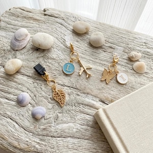 May include: Gold-toned phone charms with various pendants: a leaf, a butterfly, and a plane. Each charm has a clip for attachment. The charms also feature a light blue circle with the letter "L" and a white circle with the letter "I".