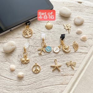 May include: A collection of gold-colored phone charms with various designs, including a tree, cat, airplane, peace sign, starfish, dragonfly, and a moon with a heart. The charms are displayed on a light-colored wooden surface with small seashells and stones.