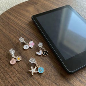 May include: Four decorative charms with various designs, including a daisy, starfish, heart, and a pink flower. Each charm has a different initial: E, J, K, and S. The charms are attached to clear clips, and are on a wooden surface next to a black tablet.