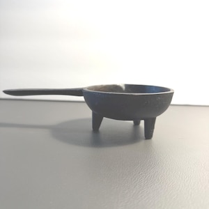 May include: A small, black cast iron pan with a long handle and three short legs. The pan is round with a slightly raised rim. The handle extends horizontally from the pan's side.