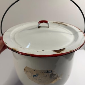 May include: White enamelware pot with a lid and a black wire handle. The rim is trimmed in red, and the lid has a red handle. A faded sticker is on the side. This vintage pot is suitable for kitchen decor.