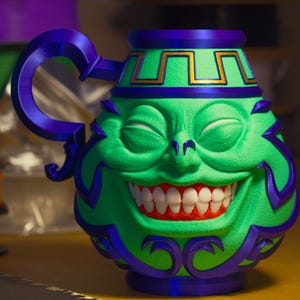 May include: A green mug with a smiling face design, featuring a wide grin with white teeth and red gums. The mug has a purple handle and accents, with gold trim around the top. The design is whimsical and detailed.