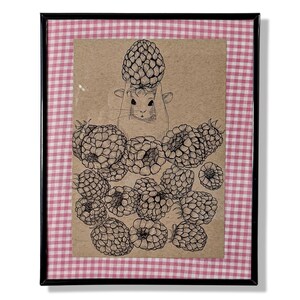 May include: Framed artwork featuring a pen and ink illustration of a small animal holding a pinecone above its head, surrounded by raspberries. The artwork is on a tan background, framed with a black frame and a pink and white checkered fabric.