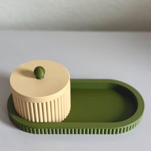 May include: A decorative set featuring a round, beige container with vertical ridges and a small green knob, placed on a green oval tray. The tray has a textured, ridged border. The set is designed in a minimalist style.