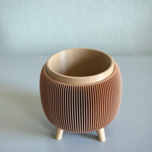 May include: A decorative, small, peach-colored planter with a ribbed exterior and a light tan interior. The planter has three short, rounded legs. The top rim and interior are a lighter shade than the exterior. The planter is empty.