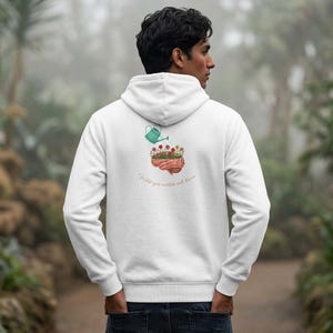 May include: White hoodie with a graphic on the back featuring a brain with flowers being watered by a green watering can. The text "What you nurture will bloom" is below the graphic. The hoodie has a drawstring hood and a kangaroo pocket.