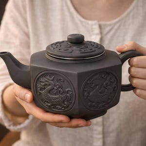 Może przedstawiać: Ciemnoszara ceramiczna czajniczek z sześciokątnym korpusem i pokrywką. Czajniczek posiada wytłaczane wzory smoków i ptaków po bokach. Uchwyt i dzióbek są zintegrowane. Czajniczek jest trzymany przez osobę.