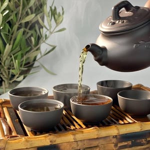 Può includere: Una teiera in ceramica marrone versa il tè in piccole ciotole abbinate, disposte su un vassoio di bambù. Il vapore sale dal tè, suggerendo che sia caldo. La scena è ambientata su uno sfondo morbido e neutro, con uno scorcio di fogliame verde.