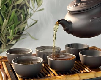May include: A brown ceramic teapot pouring tea into matching small bowls, arranged on a bamboo tray. Steam rises from the tea, suggesting it is hot. The scene is set against a soft, neutral background, with a glimpse of green foliage.