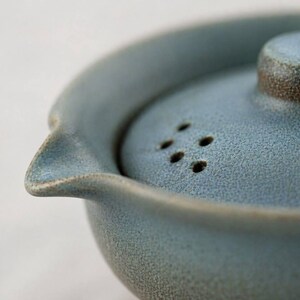 May include: Close-up of a blue ceramic teapot with a matching lid. The lid has several small holes and a brown rim. The teapot has a spout and a textured surface, suggesting a handmade or artisanal quality.