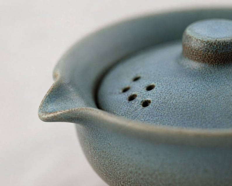 May include: Close-up of a blue ceramic teapot with a matching lid. The lid has several small holes and a brown rim. The teapot has a spout and a textured surface, suggesting a handmade or artisanal quality.
