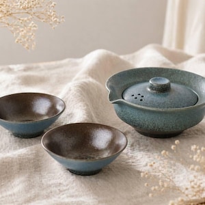 Może przedstawiać: Ceramiczny zestaw do herbaty z niebiesko-zieloną glazurą. Zestaw zawiera czajniczek z pokrywką i dzióbkiem oraz dwie małe miseczki. Miseczki mają brązowe wnętrze i niebieski zewnętrzny. Zestaw stoi na białej tkaninie.