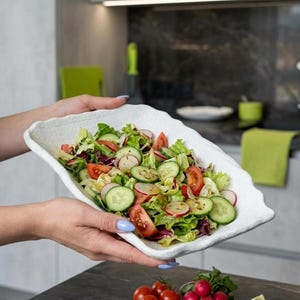 May include: A rectangular white ceramic serving dish with a textured, uneven rim filled with a fresh salad containing sliced cucumbers, red tomatoes, and radishes mixed with green lettuce leaves, presented in a kitchen setting.