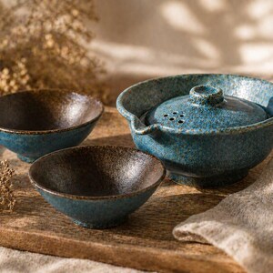 May include: A ceramic tea set with a textured, speckled blue glaze. The set includes a teapot with a lid and two small bowls. The bowls have a dark brown interior. The set is displayed on a wooden tray.