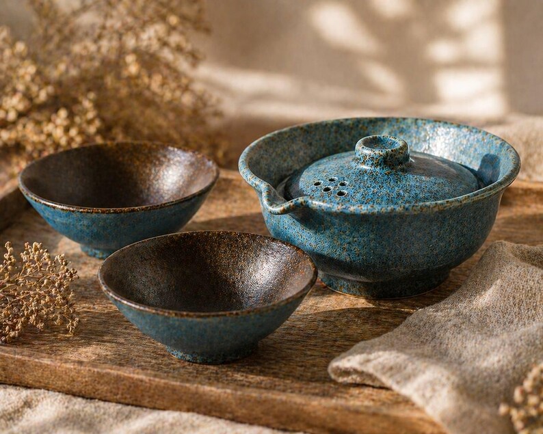 May include: A ceramic tea set with a textured, speckled blue glaze. The set includes a teapot with a lid and two small bowls. The bowls have a dark brown interior. The set is displayed on a wooden tray.
