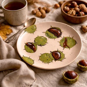 May include: A beige ceramic plate decorated with hand-painted green and brown chestnut designs. The plate is surrounded by a cup of tea, a spoon, nuts, and chestnuts in their spiky husks, evoking an autumnal theme.