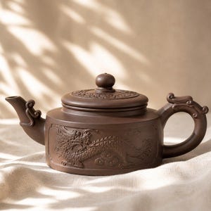 May include: Brown ceramic teapot with a dragon relief design. The teapot features a curved spout and handle, and a lid with a round knob. The dragon is carved into the side of the teapot. The teapot is sitting on a white cloth.