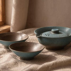 May include: A ceramic tea set with a teal and brown speckled glaze. The set includes a teapot with a lid and two small bowls. The teapot has a spout and a handle. The bowls are round and have a contrasting brown interior.