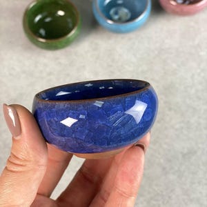 Puede incluir: Un pequeño cuenco redondo de cerámica azul con un esmalte craquelado texturizado. El cuenco tiene un borde y una base marrones. En el fondo se ven otros cuencos coloridos, como verdes, amarillos, rosas y azules.