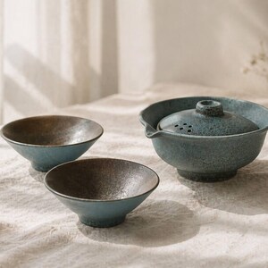 May include: A ceramic tea set with a teal and brown color scheme. The set includes a teapot with a lid and two small bowls. The teapot has a spout and a handle. The bowls are round and have a dark interior.