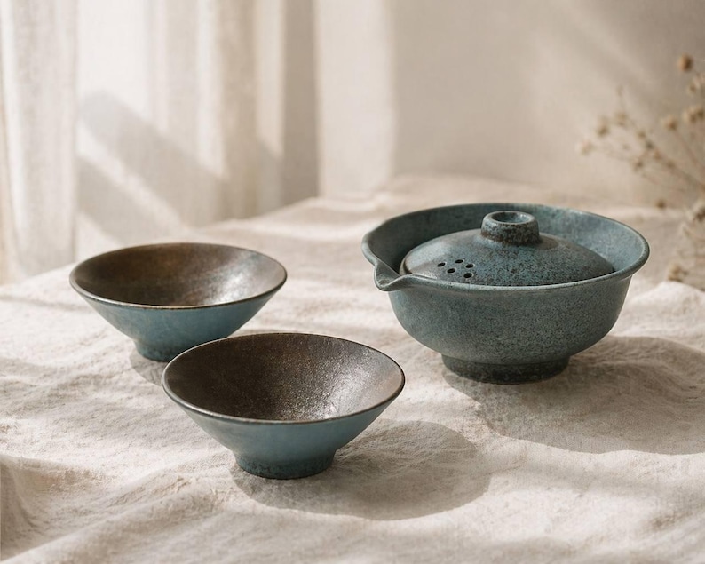 May include: A ceramic tea set with a teal and brown color scheme. The set includes a teapot with a lid and two small bowls. The teapot has a spout and a handle. The bowls are round and have a dark interior.