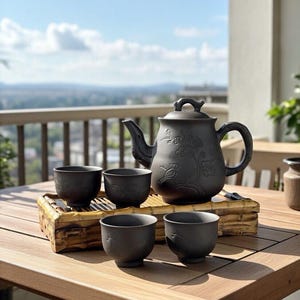 May include: A black ceramic tea set with a teapot and four cups, arranged on a bamboo tray. The teapot features floral and bird designs. The set is placed on a wooden table, with a scenic outdoor background.