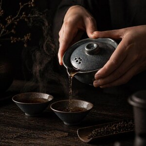 May include: A person pouring tea from a gray ceramic teapot into two small black bowls. Steam rises from the tea, and a wooden tray with tea leaves is visible. The scene is set on a dark wooden surface.