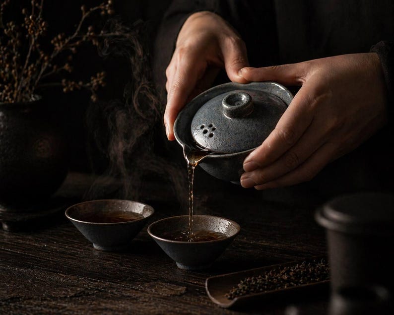 May include: A person pouring tea from a gray ceramic teapot into two small black bowls. Steam rises from the tea, and a wooden tray with tea leaves is visible. The scene is set on a dark wooden surface.