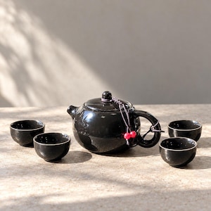Puede incluir: Una tetera de cerámica negra brillante con tapa a juego y cuatro tazas pequeñas a juego. La tetera presenta un cordón de color púrpura oscuro atado alrededor del asa y la tapa, acentuado con dos pequeñas cuentas esféricas de color rojo.