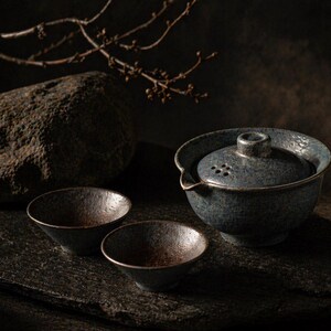May include: A dark, moody still life featuring a blue ceramic tea set with two small bowls. The tea set includes a lidded teapot with a spout and small holes. The bowls and teapot have a textured, speckled finish. The background includes rocks and a branch.