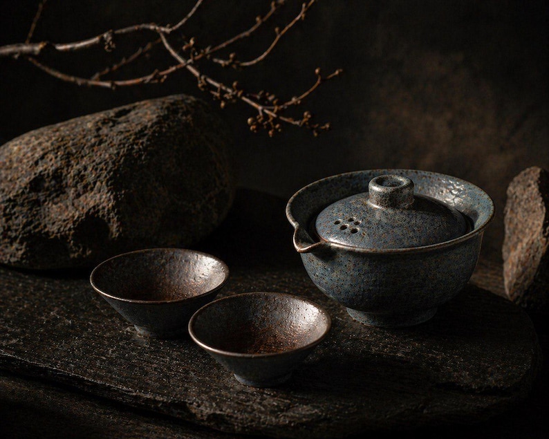 May include: A dark, moody still life featuring a blue ceramic tea set with two small bowls. The tea set includes a lidded teapot with a spout and small holes. The bowls and teapot have a textured, speckled finish. The background includes rocks and a branch.