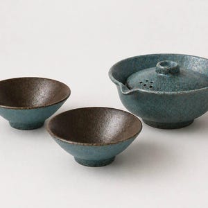 May include: A ceramic tea set with a teal and brown speckled glaze. The set includes a teapot with a lid and two small bowls. The teapot has a spout and a handle. The bowls have a rounded shape.