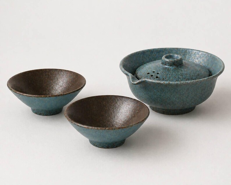 May include: A ceramic tea set with a teal and brown speckled glaze. The set includes a teapot with a lid and two small bowls. The teapot has a spout and a handle. The bowls have a rounded shape.