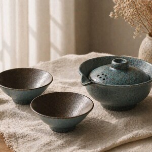 May include: A ceramic tea set with a teal and brown speckled glaze. The set includes a teapot with a lid and two small bowls. The teapot has a spout and a handle. The bowls are round and have a textured interior.