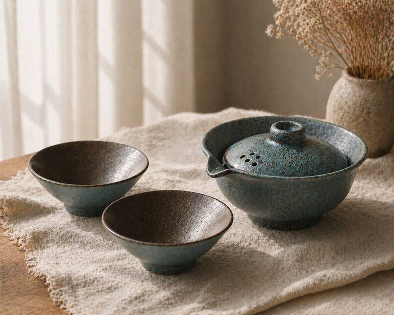 May include: A ceramic tea set with a teal and brown speckled glaze. The set includes a teapot with a lid and two small bowls. The teapot has a spout and a handle. The bowls are round and have a textured interior.