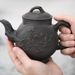 May include: A dark brown ceramic teapot with a lid, spout, and handle. The teapot is decorated with a floral and bird relief design. The teapot is held in two hands.