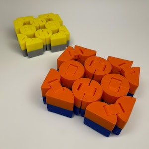 May include: Two colorful, geometric fidget toys. One is yellow with gray accents, the other is orange with blue accents. The toys are made of interlocking pieces, designed for tactile manipulation and stress relief. The shapes include cylinders and angular forms.