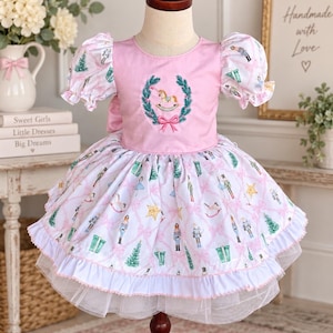May include: A pink dress featuring puff sleeves and a central embroidered wreath with a rocking horse. The skirt has a white background with a repeating pattern of nutcrackers, Christmas trees, gifts, and stars, finished with a ruffled hem.
