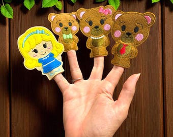 Puede incluir: Una mano que muestra cuatro marionetas de dedo de fieltro: una niña rubia con un vestido azul y tres osos de peluche marrones. Los osos tienen mejillas rosadas, y uno lleva una pajarita, un collar y una corbata roja.