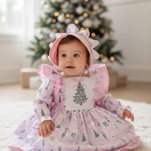May include: A baby wearing a pink Christmas dress and matching bonnet. The dress features a festive print with Christmas trees, nutcrackers, and stars. The dress has long sleeves and a square bib with a Christmas tree embroidery. The baby is sitting on a white rug.