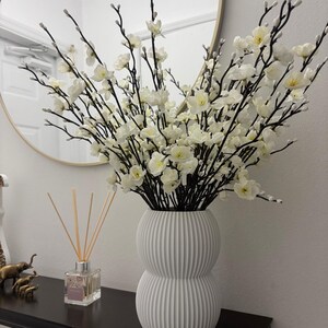 May include: A white, ribbed vase with a double-bulb shape holds a large arrangement of white flowers with black stems. The vase sits on a black shelf, with a mirror in the background. A reed diffuser and small elephant figurines are also on the shelf.