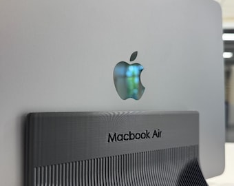 Może przedstawiać: Srebrny laptop z logo Apple umieszczony jest w ciemnoszarym, pionowym stojaku. Podstawka posiada teksturowaną, żebrowaną powierzchnię, a na środku przedniej części wyraźnie wytłoczony jest napis "Macbook Air".