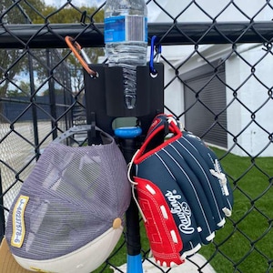 May include: A black baseball bat with a blue handle is attached to a fence, holding a water bottle, a baseball cap, and a Rawlings baseball glove. The glove is red and navy blue.