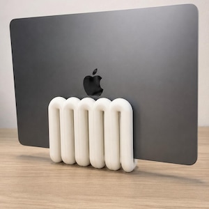 May include: A dark gray laptop with a black apple logo is held upright by a white, coiled, spring-shaped stand. The stand features a series of rounded, interconnected loops that provide a stable base for the device to rest in a vertical position.