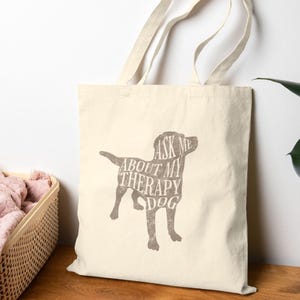 May include: Cream-colored tote bag with a brown dog silhouette design. The dog graphic contains the text "Ask Me About My Therapy Dog." The bag has two handles and is made of a canvas-like material. A basket with pink fabric is in the background.
