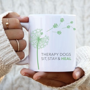 May include: White ceramic mug featuring a green dandelion design with paw prints and the text "THERAPY DOGS SIT, STAY & HEAL". The mug is held by a person wearing a white sweater.
